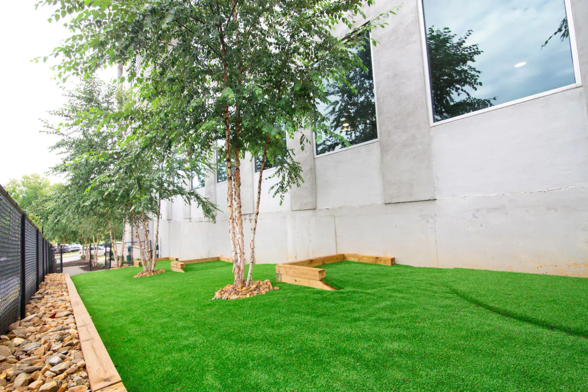 Green artificial turf with trees next to a white building. A black fence borders the left side.