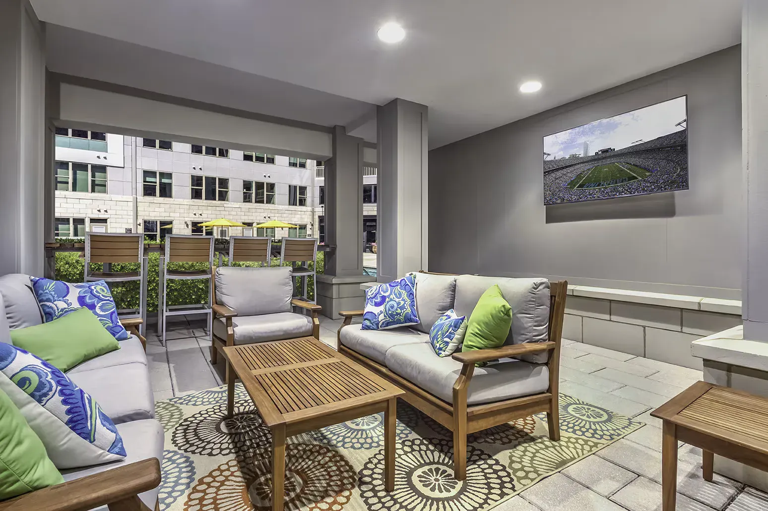 Outdoor seating area with gray and wooden furniture, patterned rug, and artwork. Buildings in background.