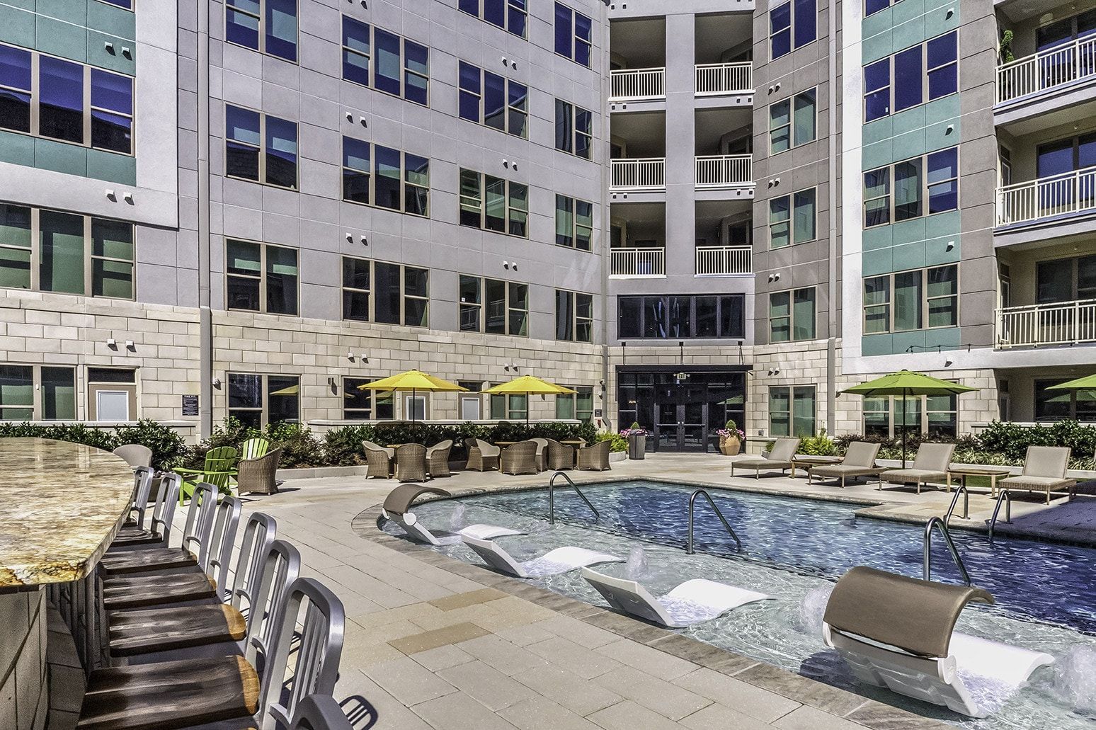 Pool area of an apartment complex, featuring lounge chairs, small tables, and several yellow umbrellas.