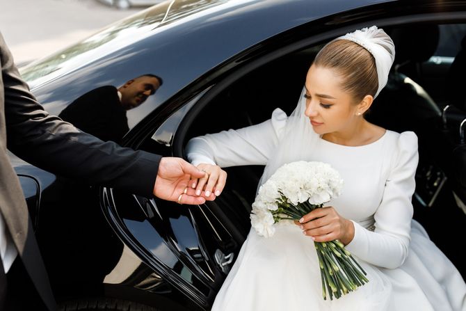 La sposa, vestita di bianco, scende da un'auto nera tenendo in mano un bouquet, mentre qualcuno le porge la mano.