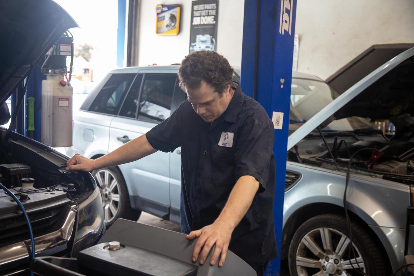 Mechanic working under the hood of a car in an auto repair shop, with another car in the background