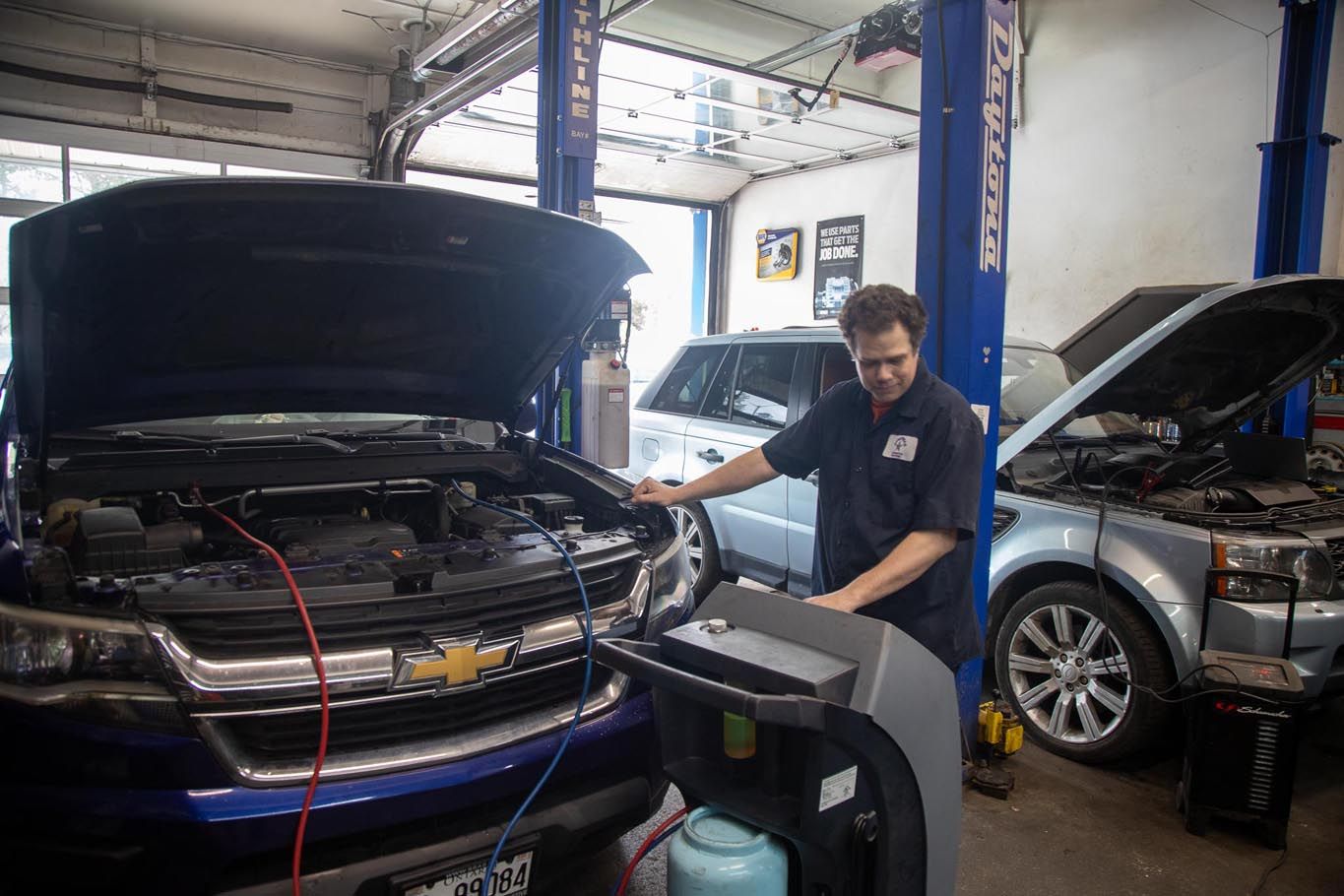 Mechanic inspecting a car engine in an auto repair shop, with another vehicle on a lift nearby