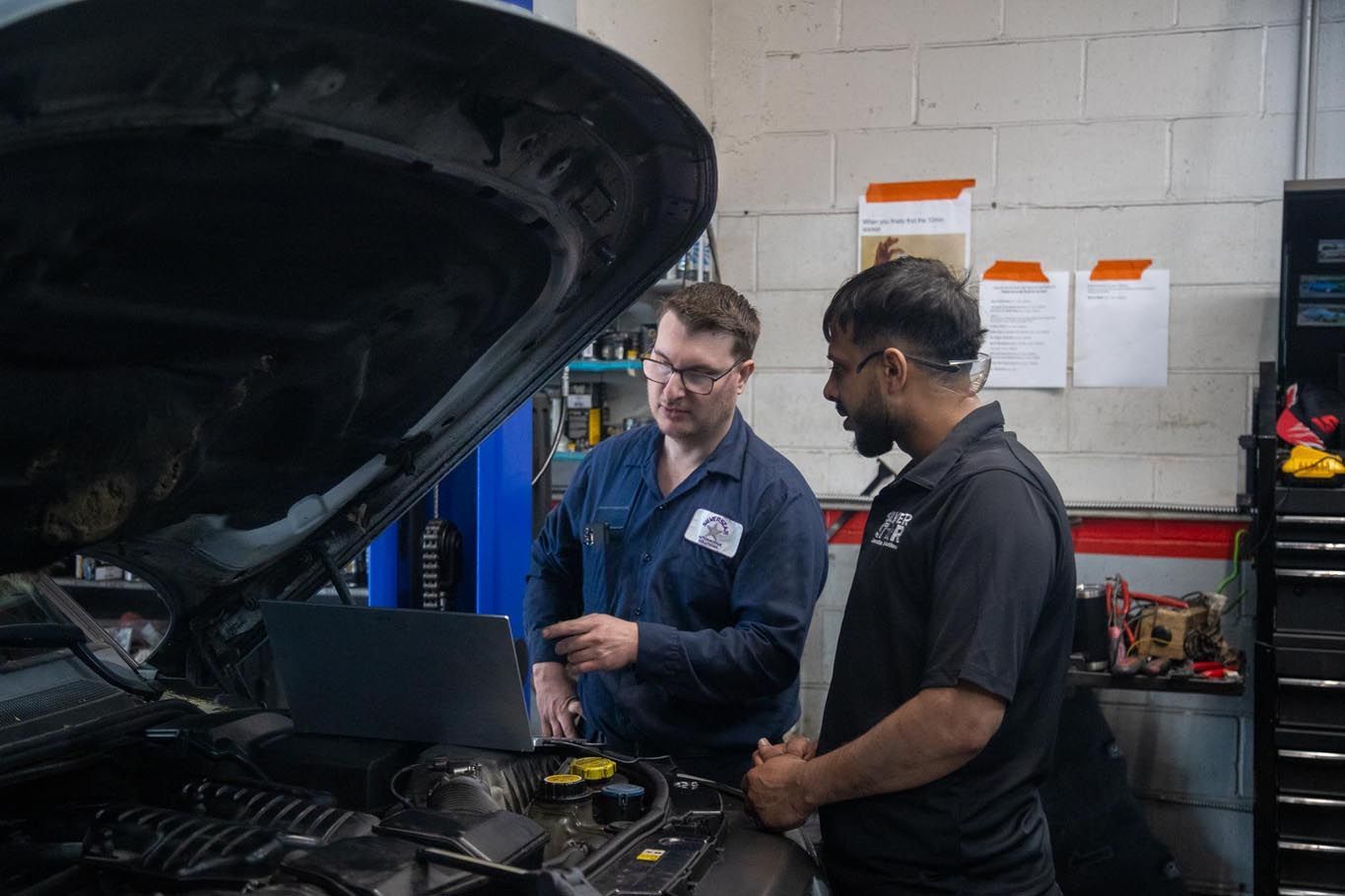 Two mechanics inspecting a car engine in a garage with the hood open