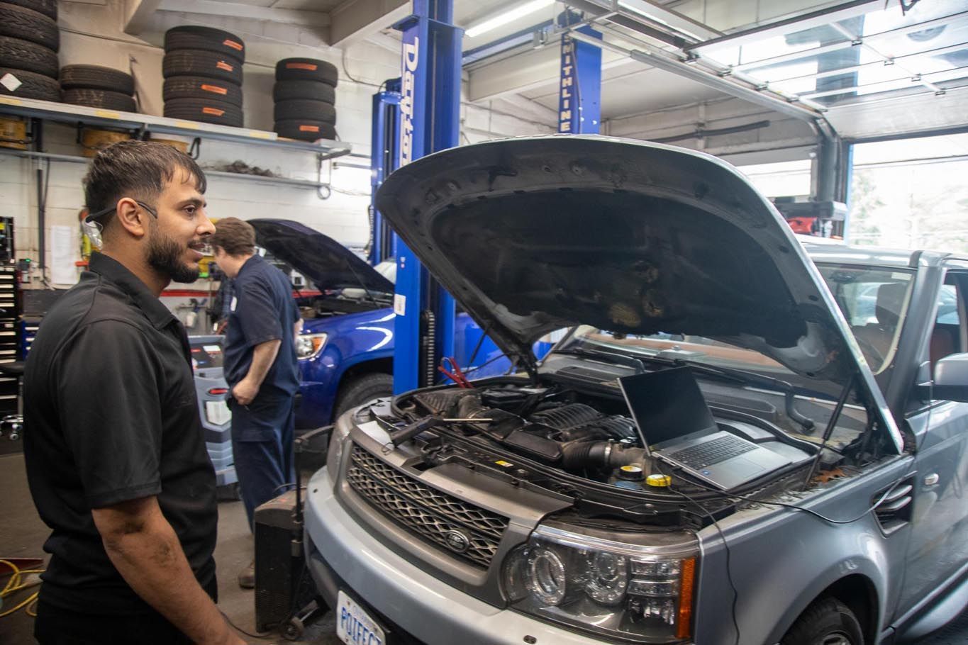 Mechanic in a garage working on a silver SUV with the hood open, tires stacked in the background
