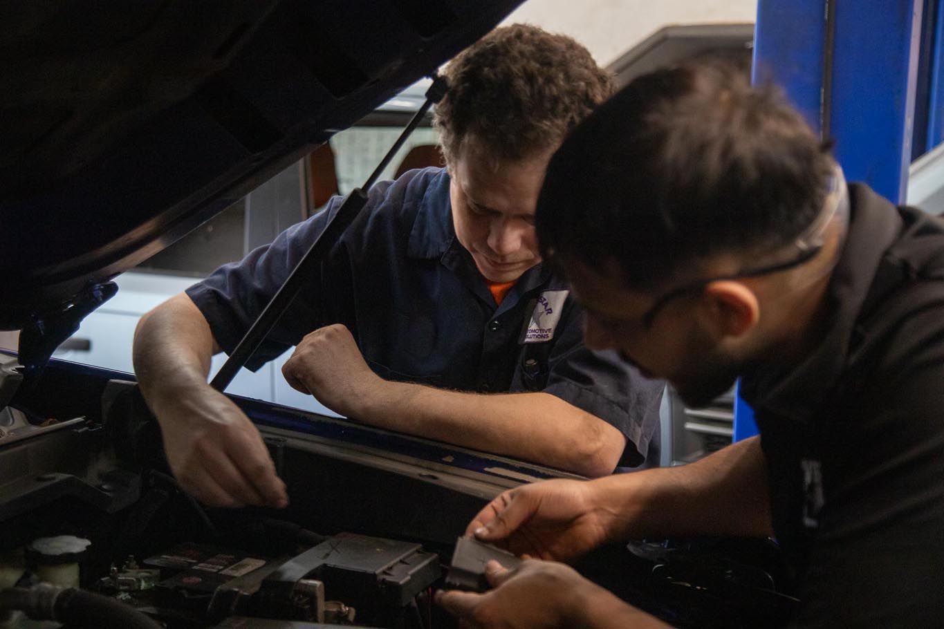 Two mechanics working under a raised car hood in a garage, inspecting the engine.