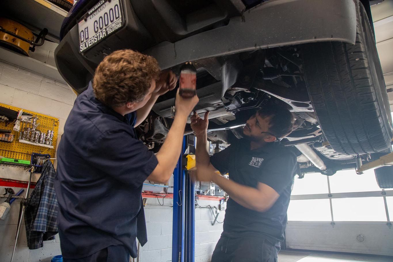 Two mechanics inspect a car underside on a lift in an auto shop