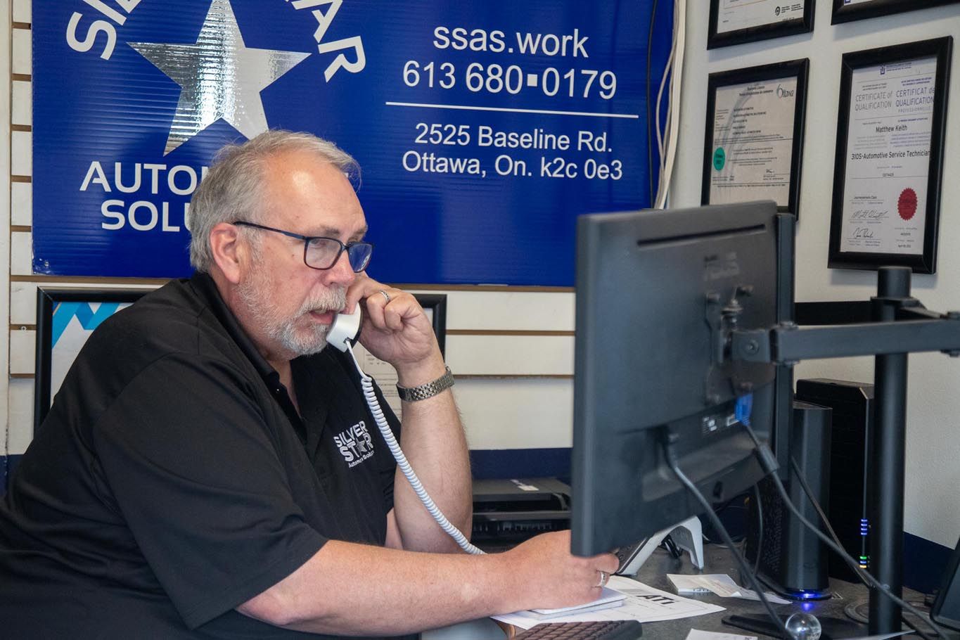 Man on a phone at a desk with a computer in an office, wearing a black shirt.