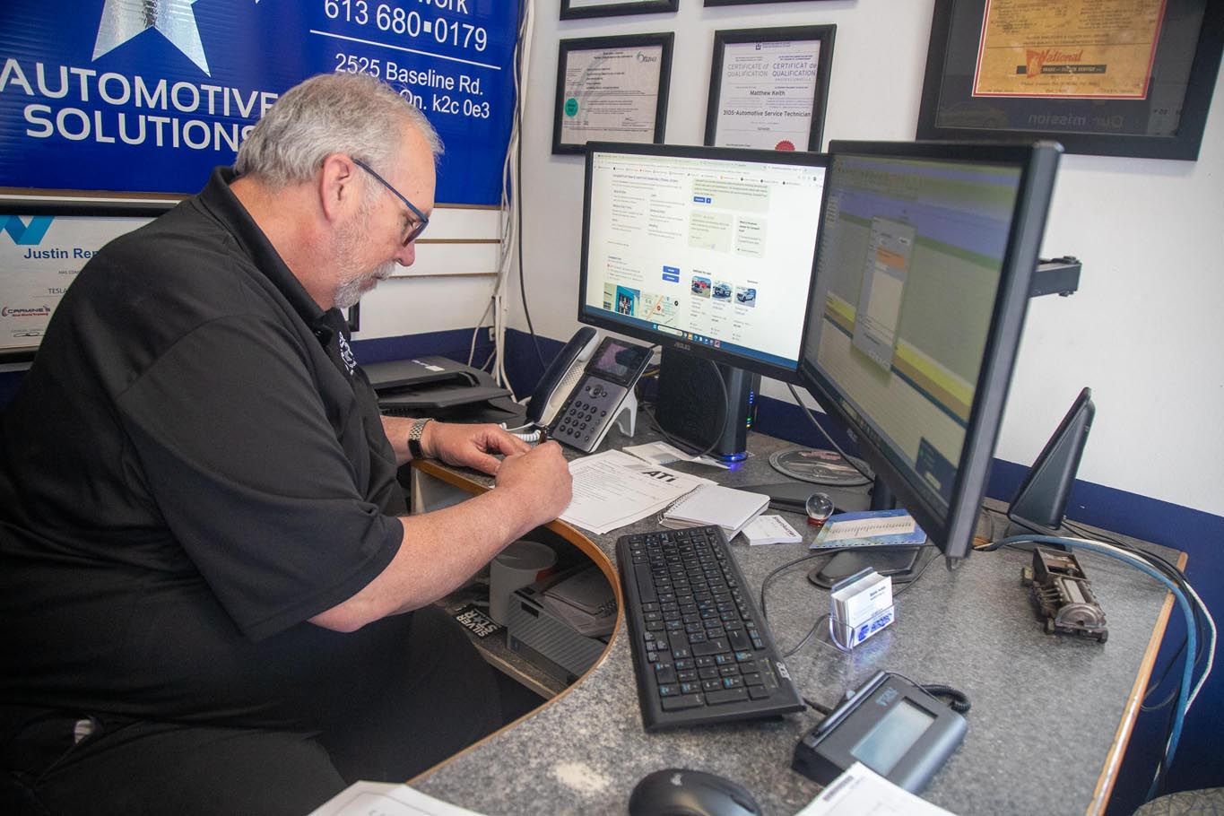 Man working at a desk with dual monitors in an office, typing on a keyboard with paperwork nearby.