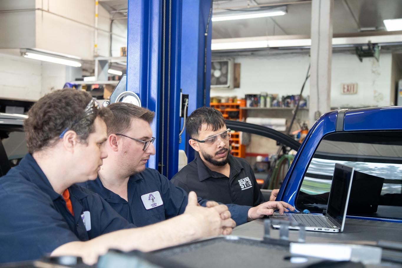 Three technicians inspect a laptop beside a blue car in an auto repair shop