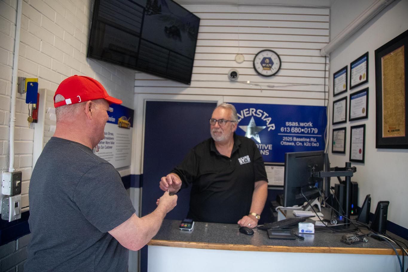 Customer and clerk shaking hands at a service counter in a small office, with a TV and framed certificates on the wall.