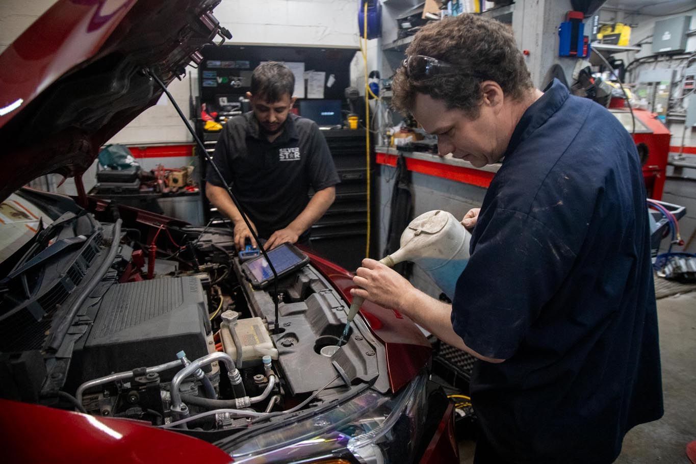 Two mechanics working under a red car hood in a garage, one holding a part while the other inspects the engine
