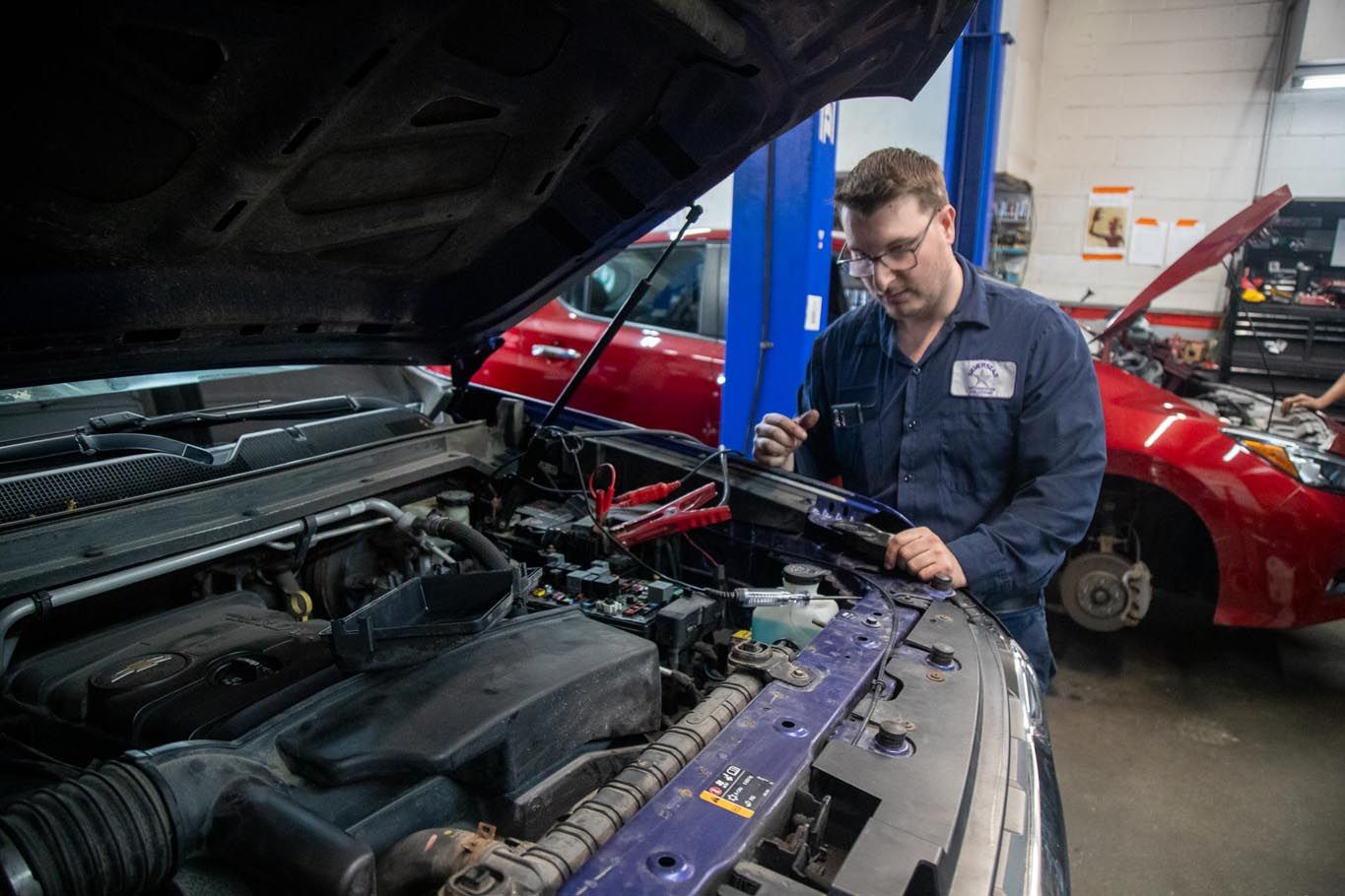 Mechanic inspecting a car engine in a repair shop, with a red car and open hood in the background