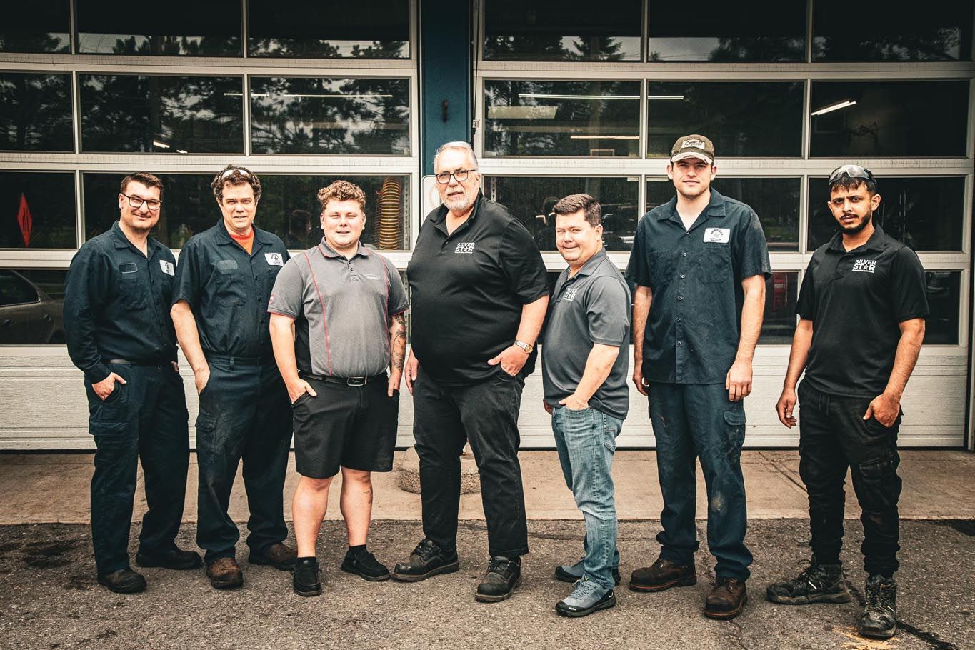 Six people standing in a row outside a garage, wearing dark work shirts and boots.