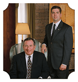 Two men in suits posing indoors, one seated, one standing, leaning on a wall.