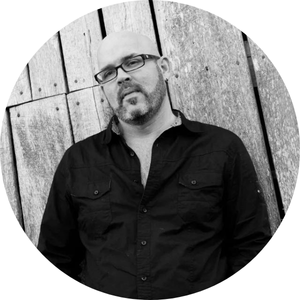Bald man with glasses wearing a black shirt leans against a wooden wall, looking slightly off-camera.