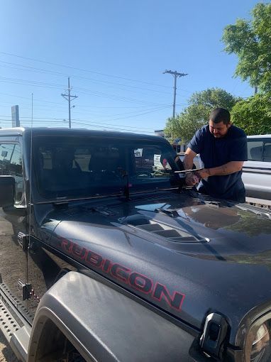 Jeep windshield replacement in Amarillo, TX Man wiping the windshield of a black Jeep Rubicon under a bright blue sky outdoors