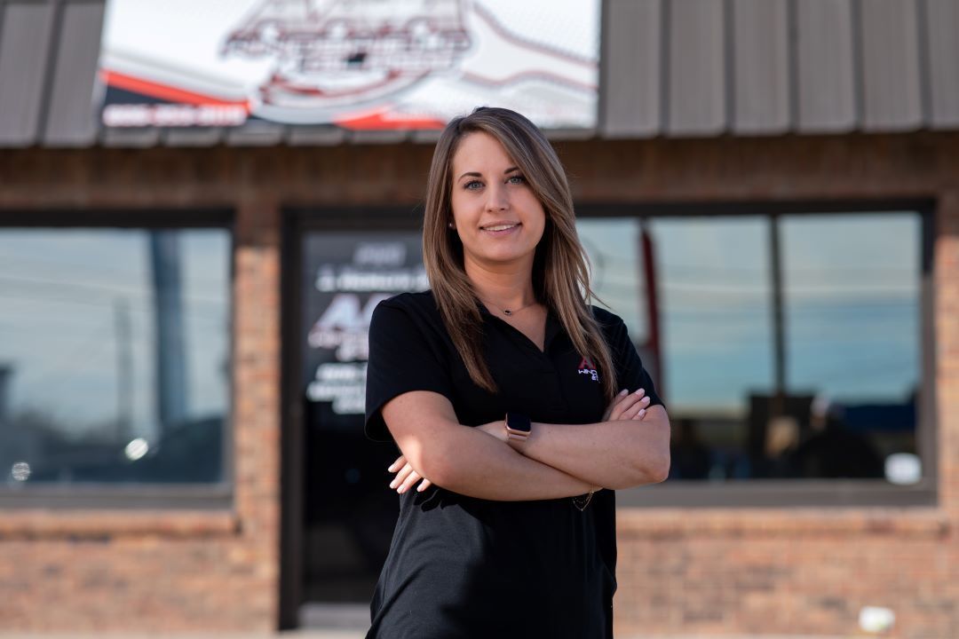 Woman standing with arms crossed in front of a brick building, wearing a black shirt and smiling.