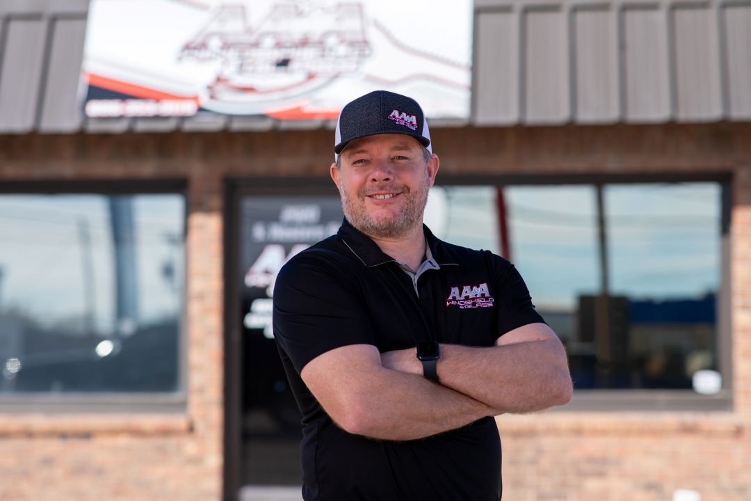 Smiling man in a black shirt and cap standing with arms crossed outside a building