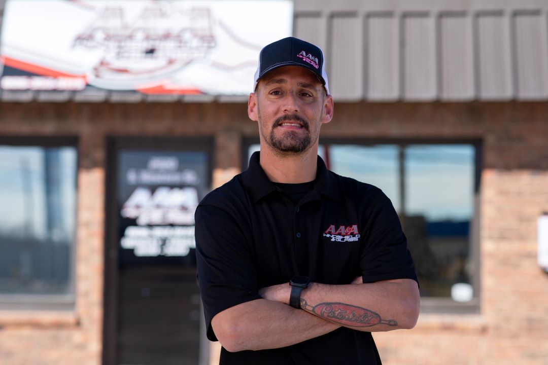 Man standing with arms crossed outside a storefront, wearing a black shirt and cap.