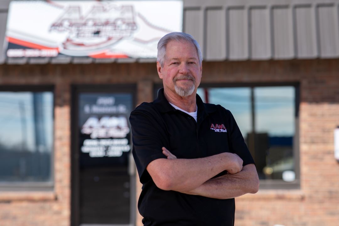 Man standing with arms crossed in front of a building entrance, wearing a black polo shirt.