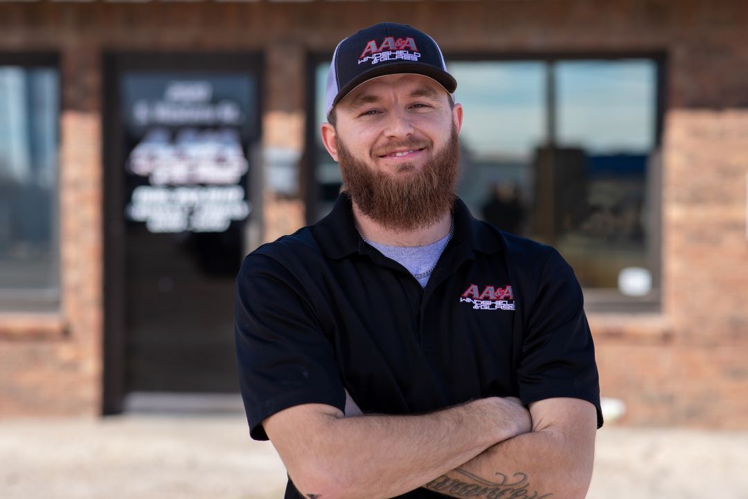 Smiling man in black A&A polo and cap standing with arms crossed outside a brick storefront