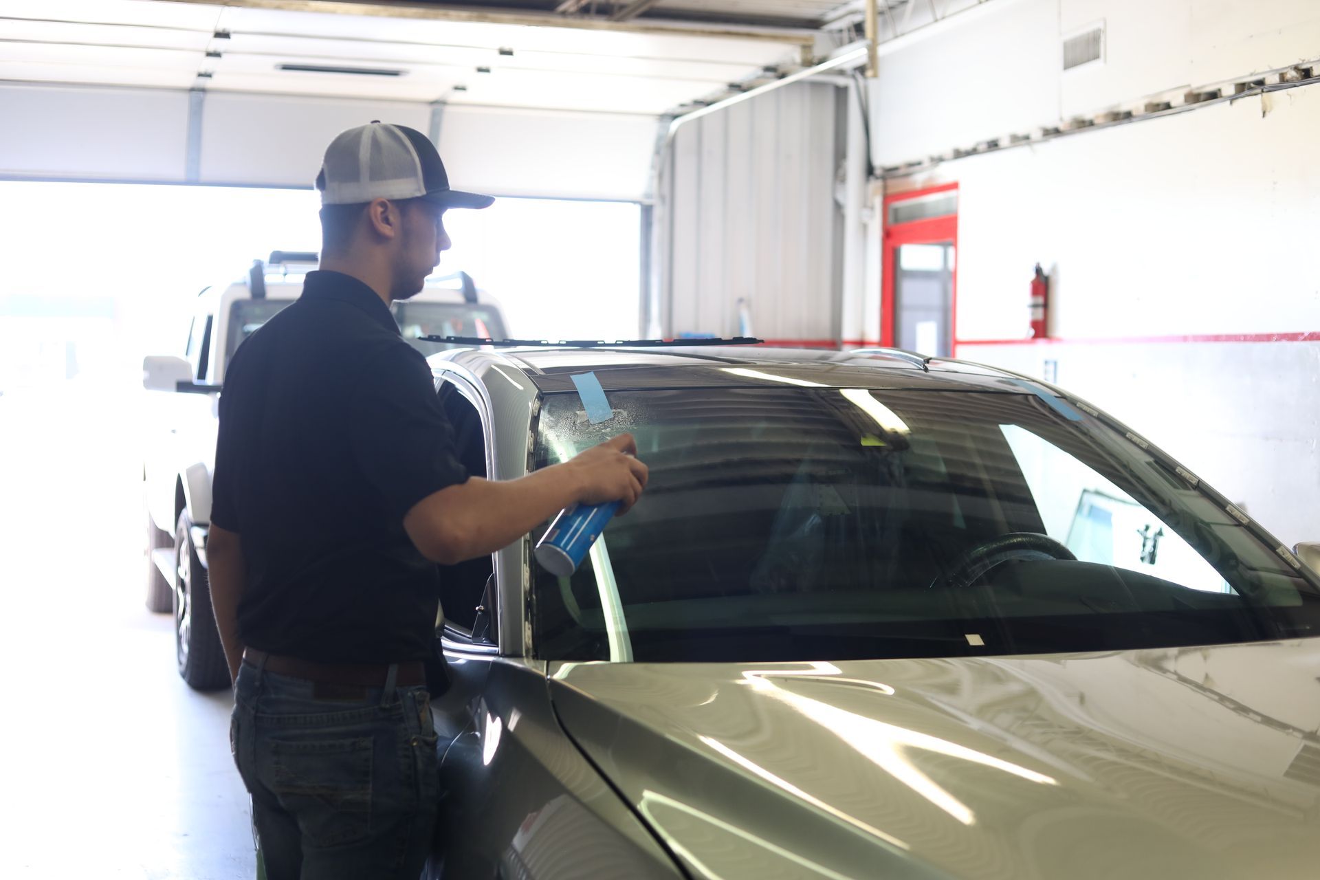 Person cleaning a silver car windshield in a bright garage with another vehicle in the background