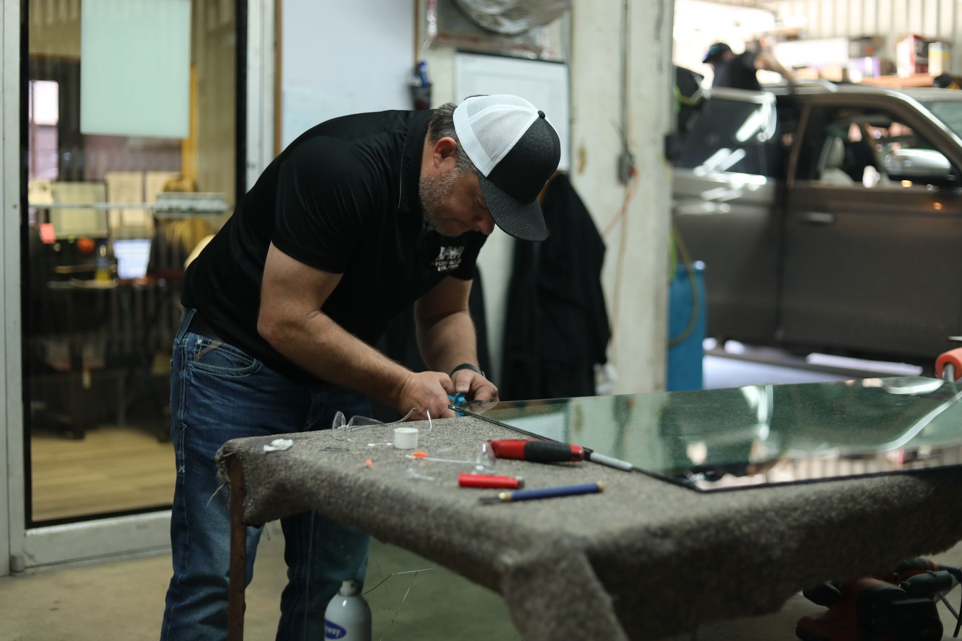 Man in a black shirt and cap working on a glass pane in a workshop, with tools on a table.