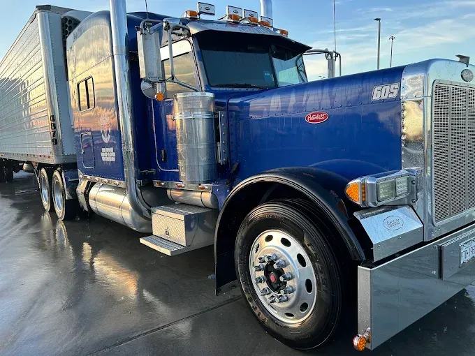 Blue semi-truck with chrome grille and trailer parked on wet pavement in a lot