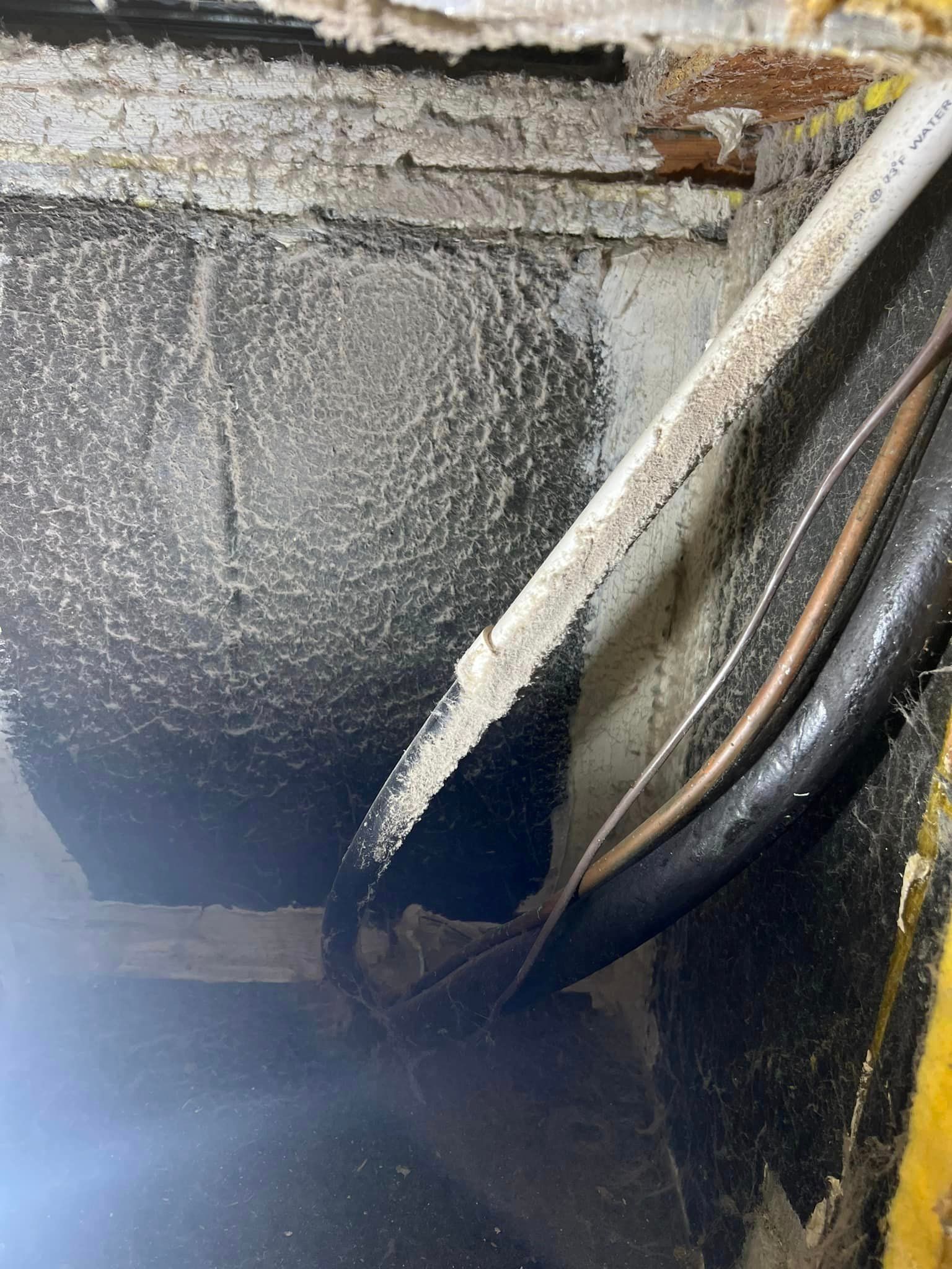 Close-up of a damaged wall cavity with exposed insulation, pipes, and peeling material around a dark opening.