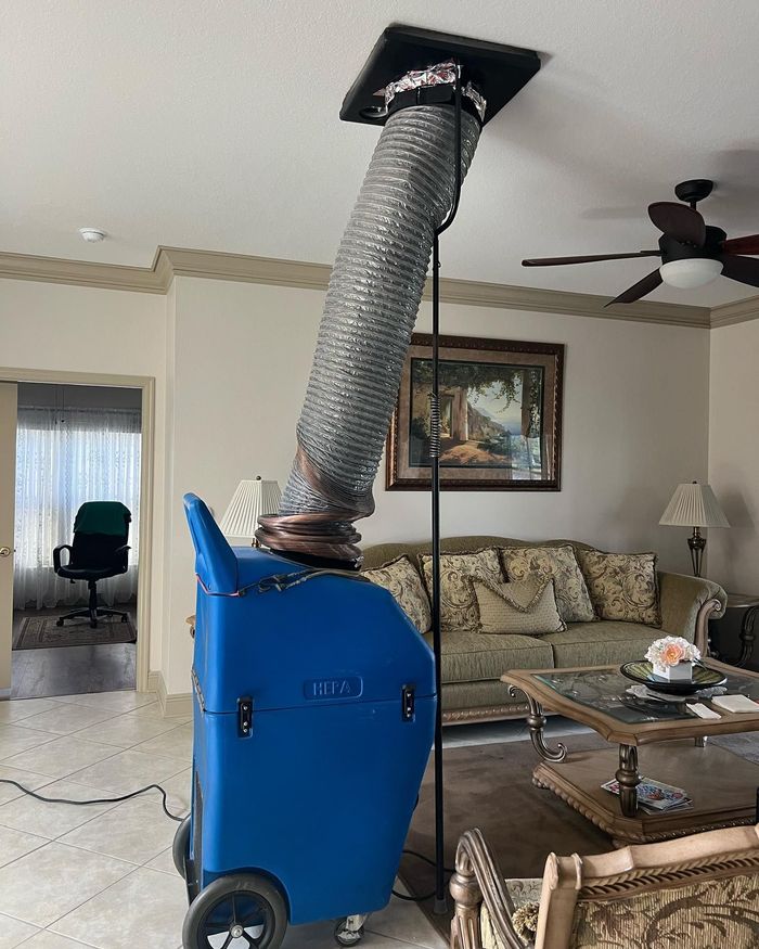 Blue portable ventilation unit with a silver flexible duct reaching to a ceiling vent in a living room