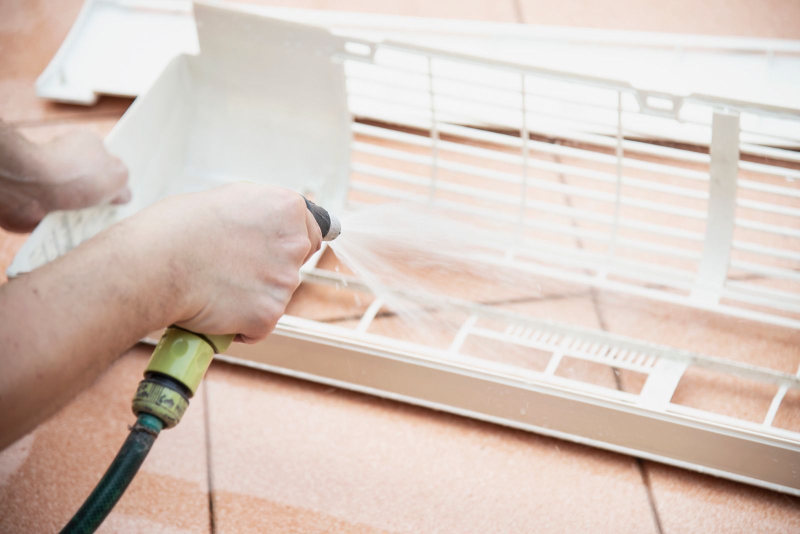 Hands using a spray nozzle to clean white window blinds by a tiled floor.
