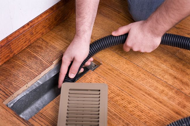 Hands using a vacuum attachment to clean dust from a hardwood floor vent.