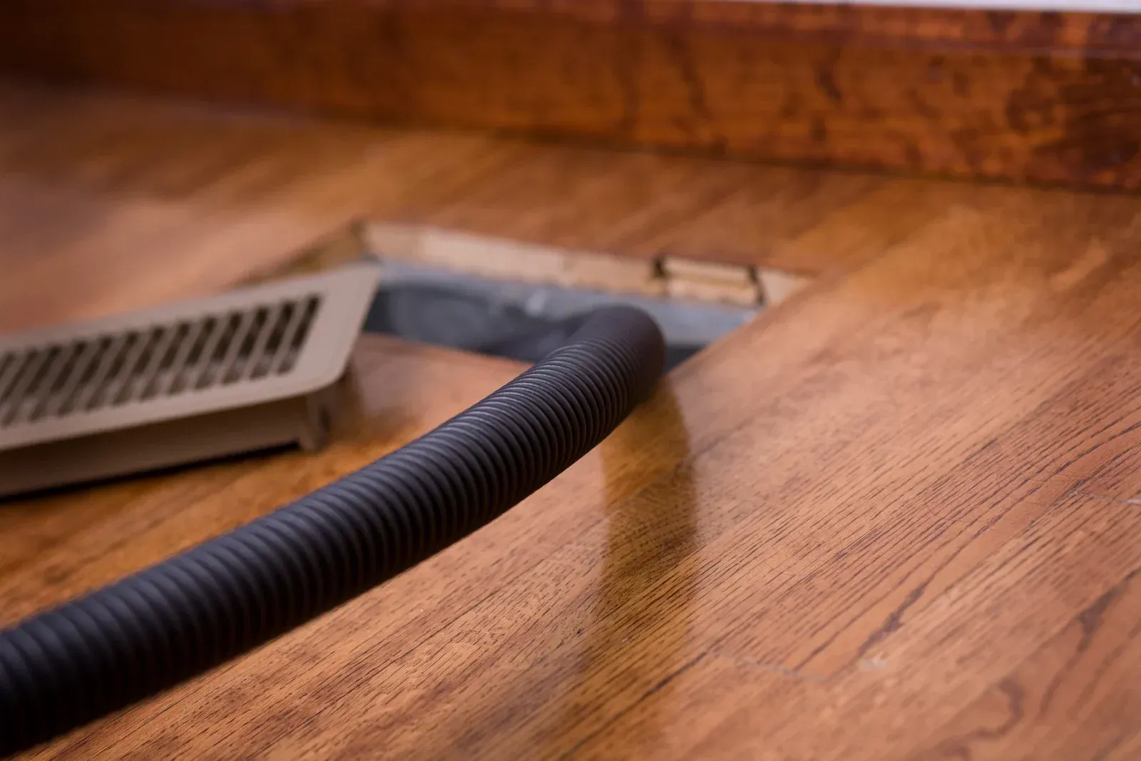 A vacuum hose cleaning a floor vent in a wooden floor.