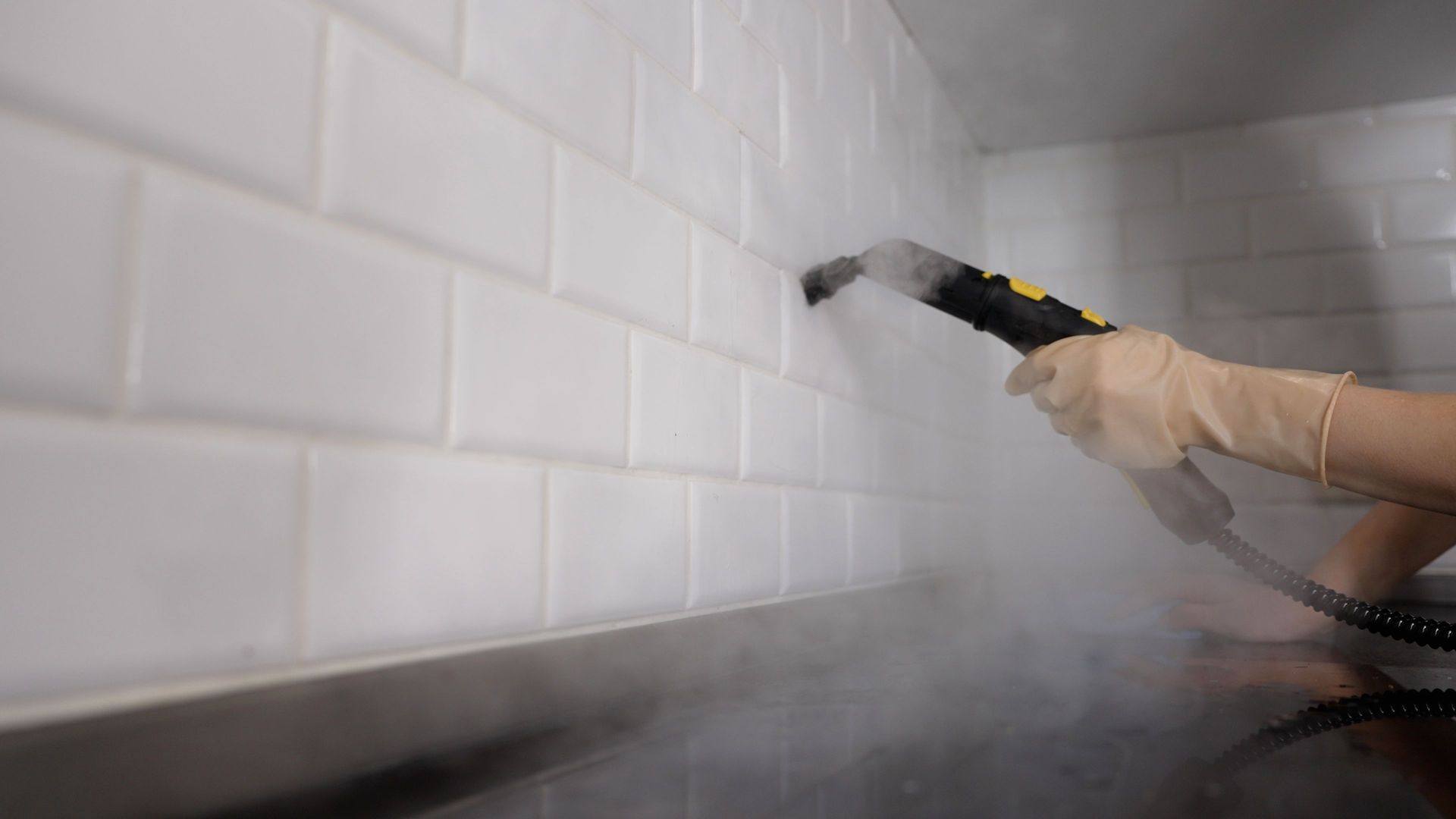 Hand holding a steam cleaner spraying a white tiled wall with visible steam.