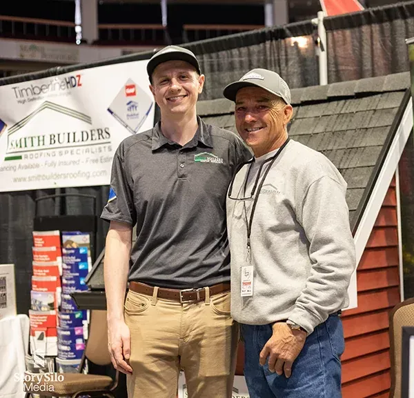 Two men smiling, posing at a booth with a roof display, Smith Builders sign, at an event.