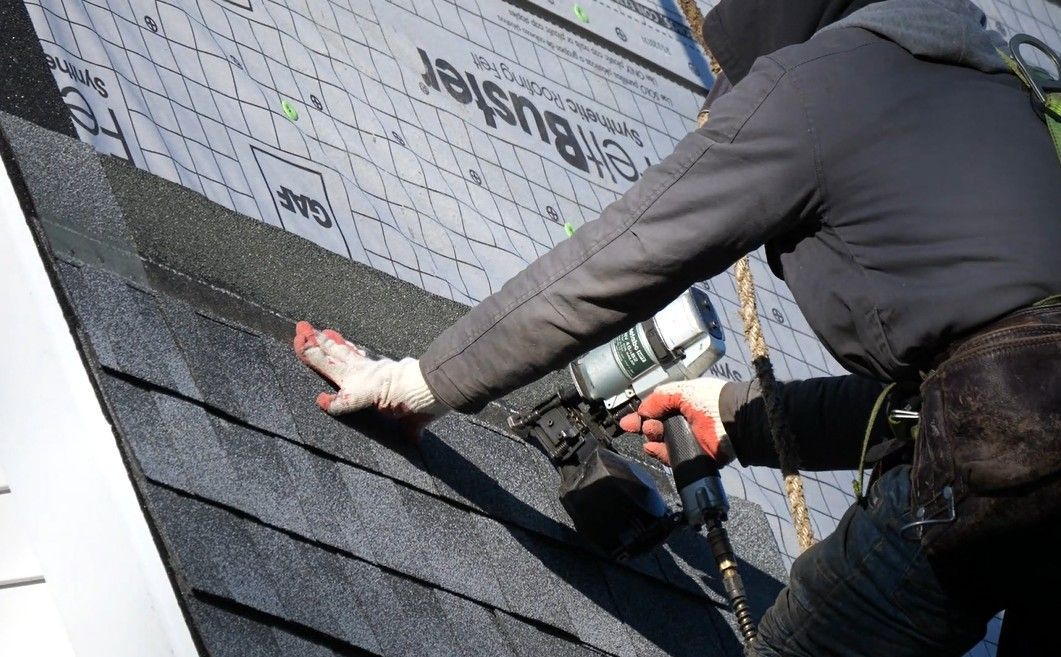 Roofer installing asphalt shingles on a roof; using a nail gun; wearing gloves and safety gear.