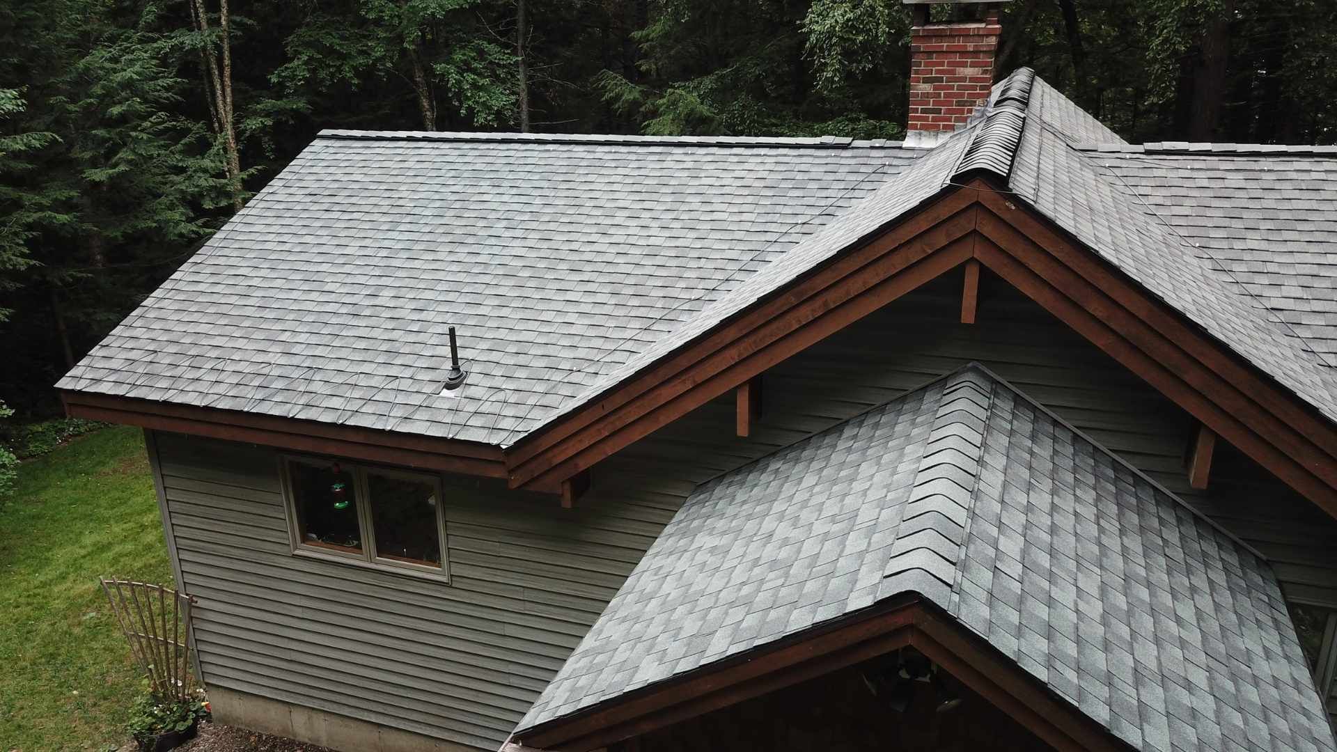 Gray shingled roof of a house with a brick chimney, set among trees.