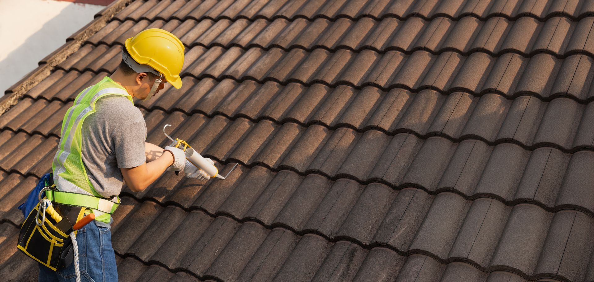 Roof worker in yellow hard hat securing brown roof tiles with a power tool