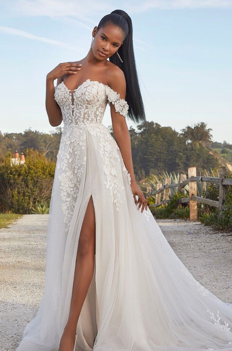 Woman in white wedding dress with lace bodice, long sleeves, and flowing skirt, posing in front of floral backdrop.