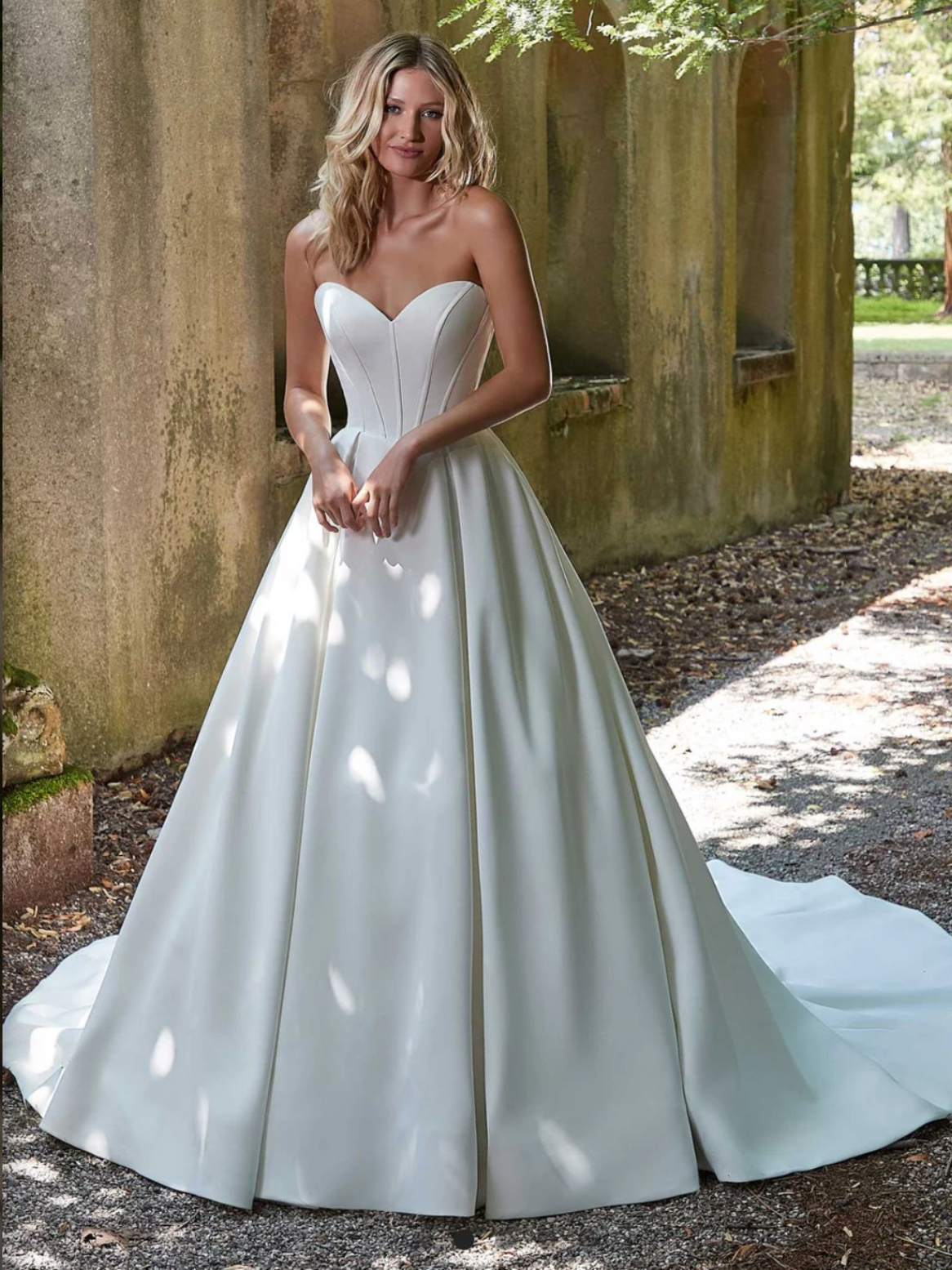 Woman in a white off-the-shoulder wedding gown with long sleeves, standing in front of floral arrangement.