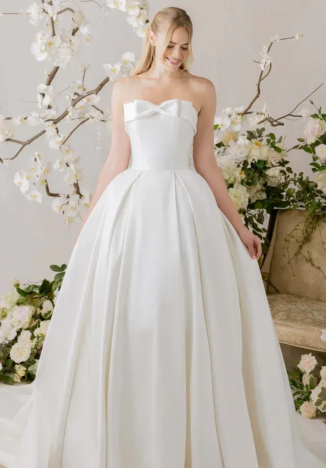 Woman in white strapless wedding gown with bow, standing, smiling, with flowers and chair in background.