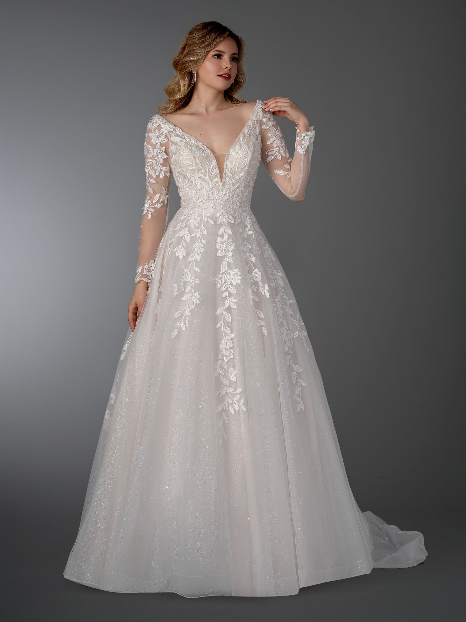 Woman in a white wedding gown with long, sheer sleeves. Deep V-neckline, floral detailing, and a flowing skirt.