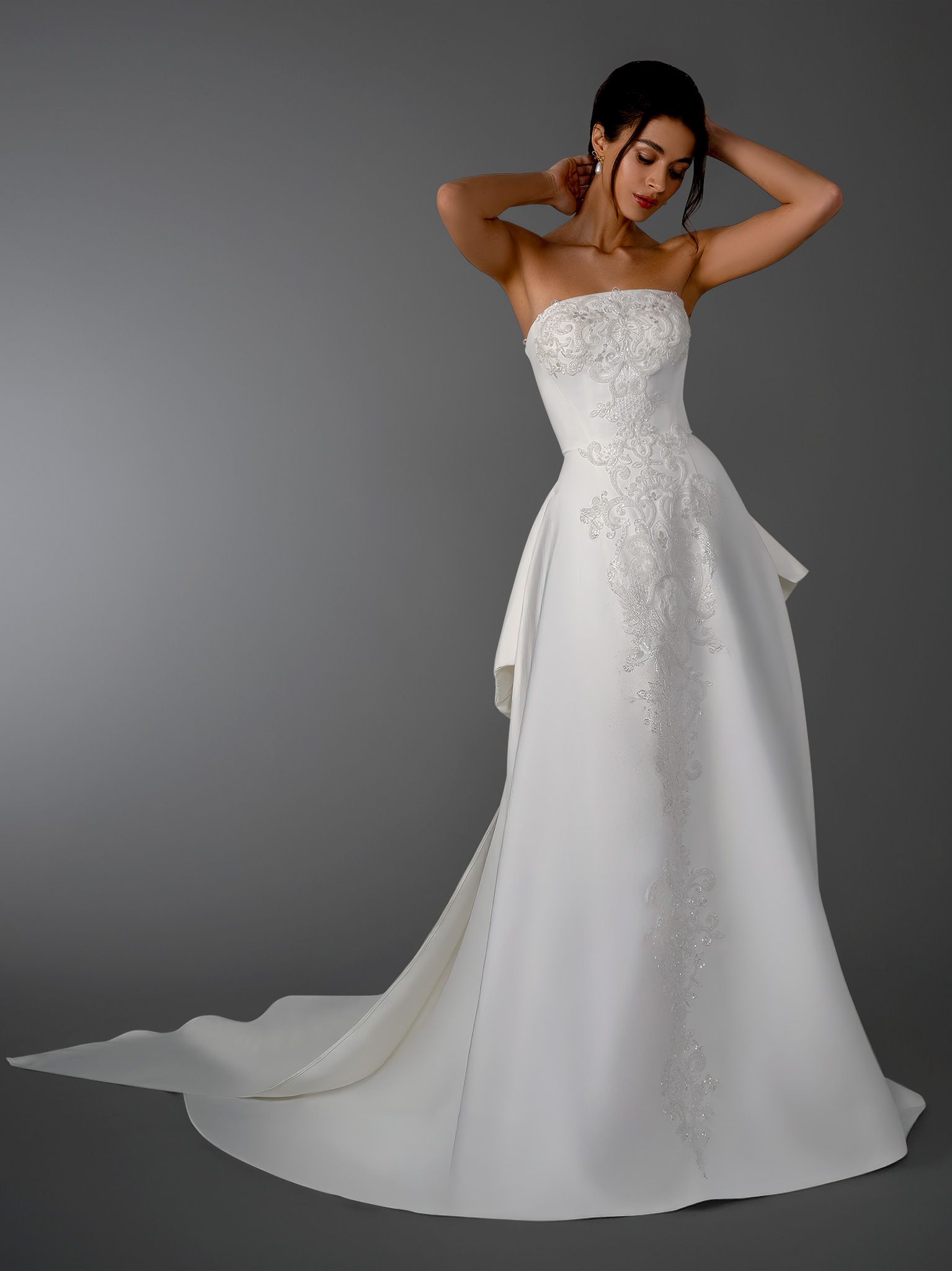 Woman in strapless white wedding dress, arms raised, against gray backdrop.
