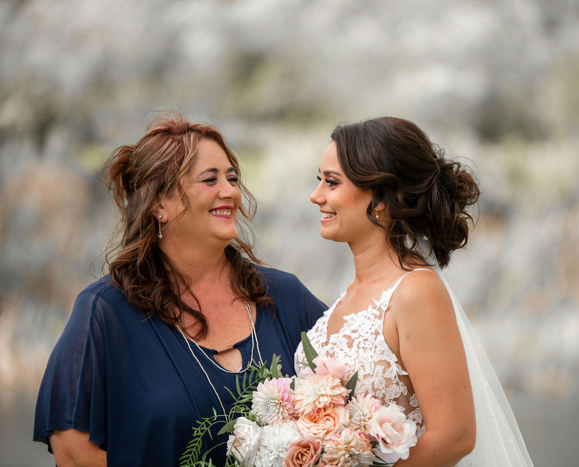 Bride smiles at woman, holding a bouquet. Woman in blue dress smiles back.