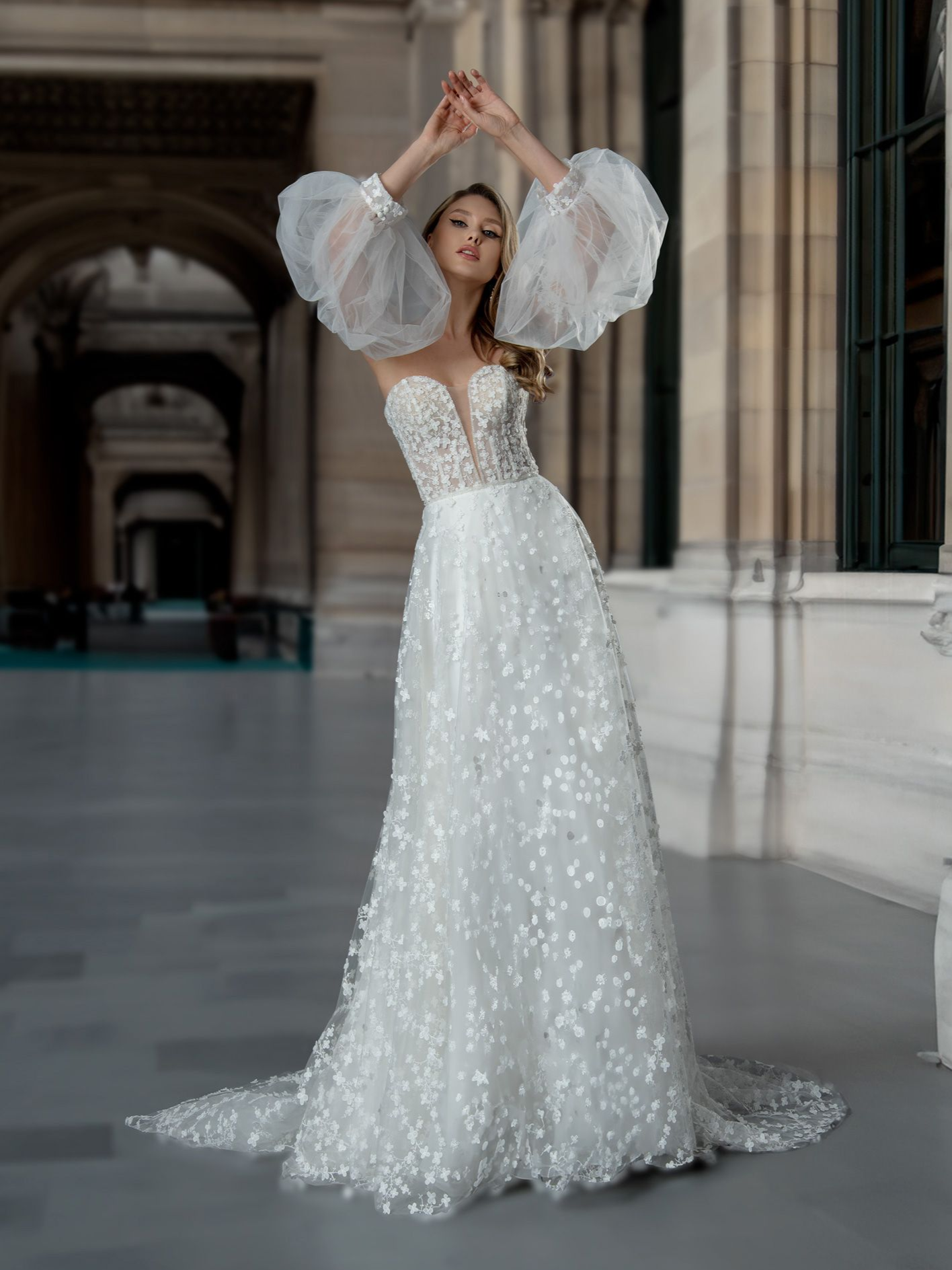 Woman in white floral gown with sheer, puffy sleeves, arms raised, in a stone hallway.