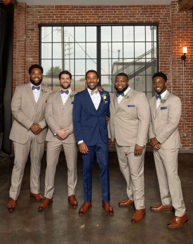 Five men in suits pose for a photo in front of a window. One man wears a blue suit; the rest wear tan.