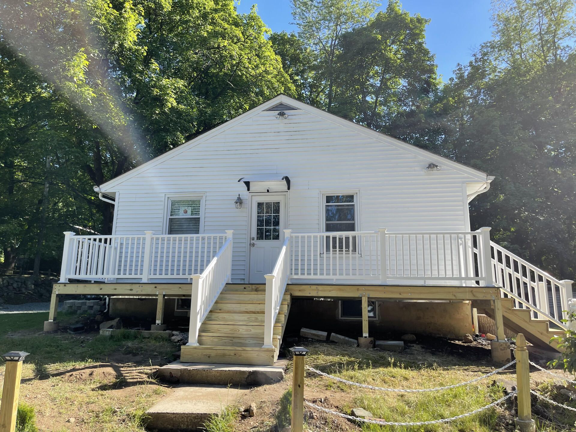 White house with a wraparound deck, steps leading to the front door, surrounded by trees.