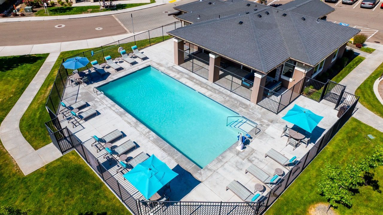 An aerial view of a fenced community pool with lounge chairs and blue umbrellas beside a clubhouse at Olympus at Ten Mile, offering townhomes in Meridian, ID.