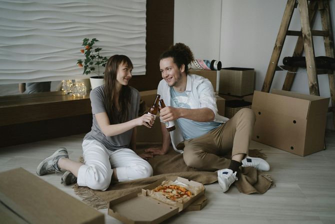 Couple toasting beers, sitting on floor surrounded by moving boxes, pizza, and ladder.