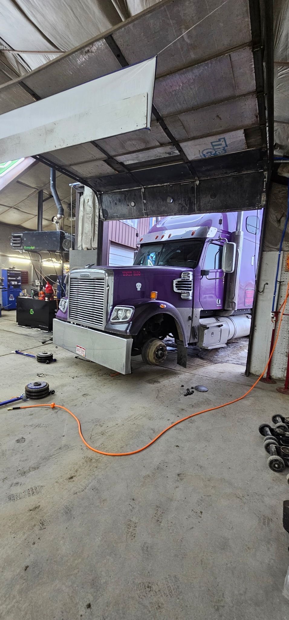 A purple semi-truck sits inside a workshop with its front wheel removed, undergoing repairs on a concrete floor.